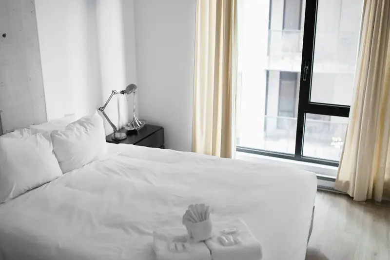 Lit d'hôtel aux draps blancs impeccables près d'une grande fenêtre, lumière naturelle du matin